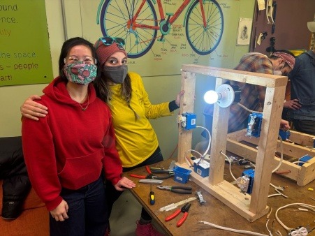Two people smile at the camera while a structure used to teach about electrical circuits with a lightbulb in it glows beside them
