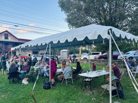 An example of Community Meal held outdoors during warmer months A white tent set up on a green lawn, with rectangular tables and chairs set up beneath its shade and a multidude of folks standing, sitting, eating, and moving around the area