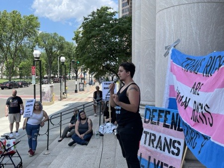 PeerPride team member speaks in front of a group of people at an outdoors trans rights community speakout. Behind him is a trans flag colored banner with the words "trans rights are human rights" and a trans flag colored sign that says "defend trans lives". 