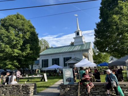 The green at Grace United Methodist Church, a community gathering location for Plainfield Farmers Market and Community Meal, amongst other community gatherings. A low stone wall encloses a green roofed, steepled, white church in the background with a number of farmers market booths and folks beneath shade tents on the right hand side and a table with food on it on the left hand side, a folding green sign states 'Plainfield Farmers Market, 4-7pm'.