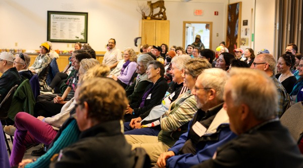 A group of 80 some people sit in rows indoors looking forward and smiling. Ages vary from people in their 20s to people in their 80s. 
