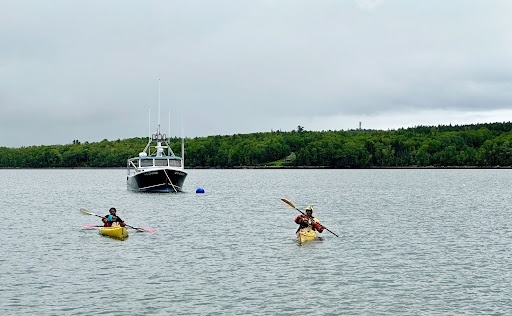 Two kayaks paddle towards the viewer, with a bigger boat appearing to safeguard them from behind. Photo credit: Deb Parker