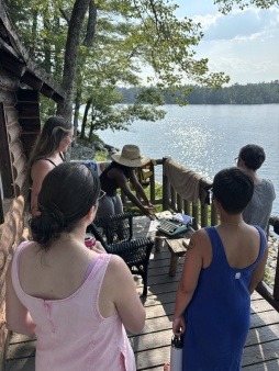 Liz Rhaney, a black artist, stands on a deck in front of a beautiful lake, talking about her artwork to a group of fellow artist residents in 2025