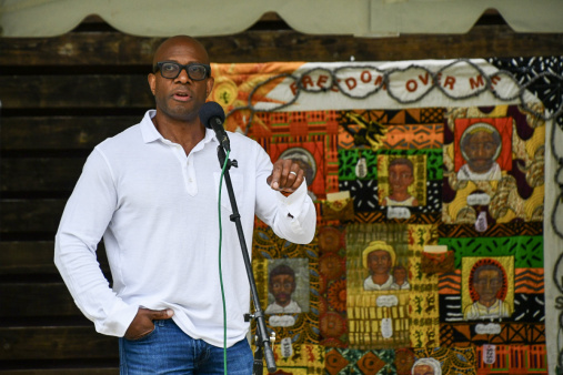 Person of color, wearing a white shirt and black glasses, speaks in a microphone. Backdrop is a colorful quilt.
