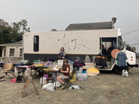 The Joy Truck is parked in a parking lot at the Milo Flea and Farmers Market. It is a smoky day from Canada's wild fires and one of th Joy Truck team members stands holding an armful of clothes while a young artist draws at the corner table surrounded by donated clothes, toys and shoes. 