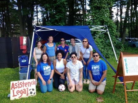 Group of people sitting and standing under a sunshade in the summertime beside a "Legal Assistance" sign in Spanish