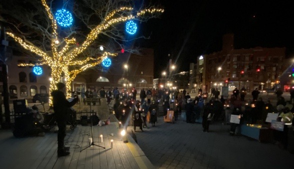 A speaker stands on a stage in an outdoor park while many people look on holding candles at a vigil