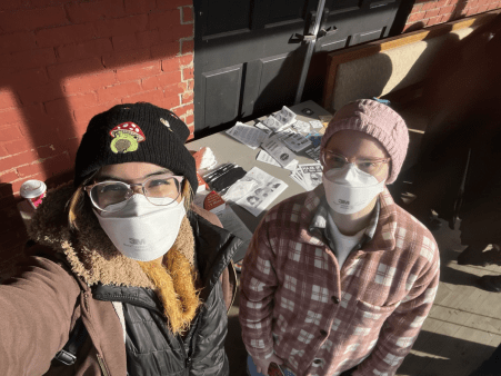 Two Clean Air Club BTV Members pose in masks in front of their table at the Barre Queer Craft Fair
