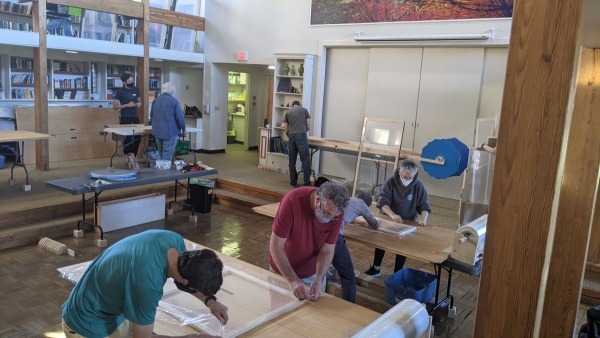 Several people work in pairs or individually at many tables attaching plastic film to wood window insert frames, attaching foam around the edges, or building the frames themselves. The room is a two storey high school library.