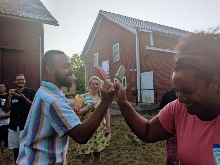 Two people tapping popsicles in an outdoor setting.  They are smiling and laughing.  Behind them is a big, red barn and other people smiling.