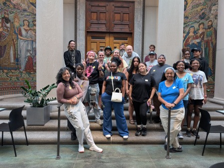 Group of people from many backgrounds and identities posed in front of a mosaic mural 