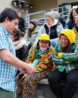 a group of people sit at an outdoor amphitheater. Two people bundled up in bright neon colors sitting side by side smile down at a turtle held firmly in a wildlife rehabilitation worker's hands. 