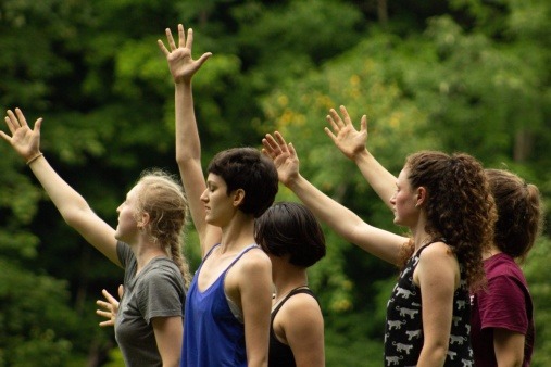 Five dancers stand outdoors, facing to the right, with their right arms stretched out in front of them. 