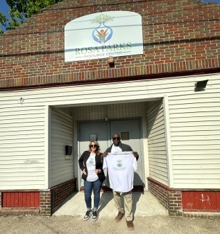 Arthur Johnson and Bernice Morris standing in front of the Rosa Parks Resource Center. Bernice is white woman with brown shoulder length hair, eye glasses,  a brown jacket with a white t shirt with the Rosa Parks Resource Center on it. Arthur is a tall Black man with a low haircut, sunglasses, wearing a leather jacket, holding a white Rosa Parks Resources Center t shirt, wearing tan pants
