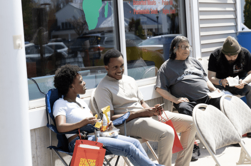 A group of people are seated outdoors in front of a building on a sunny day. A young Black woman and a young Black man are in the foreground, talking and smiling. The woman holds a bag of chips. Two other people, a person with gray hair and glasses and a man with a beard and beanie, are seated next to them. The man is looking at his phone or a book.