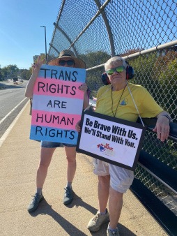 Two New Hampshire Bridge Brigade for Democracy volunteers stand on an overpass holding supportive signs, including one reading “Trans Rights Are Human Rights” and another saying “Be Brave With Us. We’ll Stand With You.” They are participating in a peaceful, community-led visibility action promoting safety, inclusion, and solidarity.