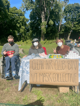 Three members of Clean Air Club BTV and Vermont Mask Collective sit at a table in a field on a sunny day. There is a sign that says Clean Air Club / VT Mask Collective. All are wearing masks and one person sits in a wheelchair. 