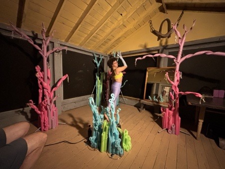 Viva, a black performance artist, is pictured in a Hewnoaks' cabin, dancing among colorful sculptures.