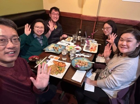 Five Asian Americans sitting around a table at sushi restaurant, food on plates in front of each person. Each person is smiling and waving.