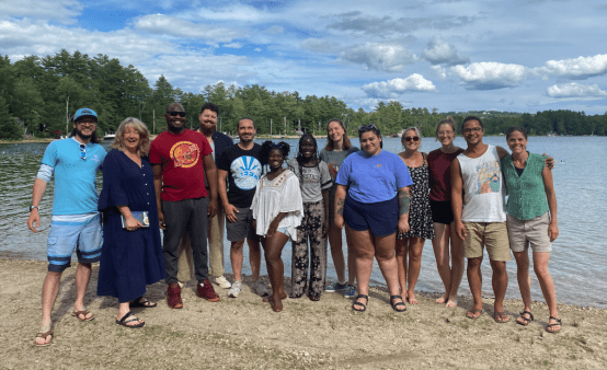 A photo showing 13 people standing together on sand by a lake and trees in the background. The people are of different races, ages, heights, sizes. 